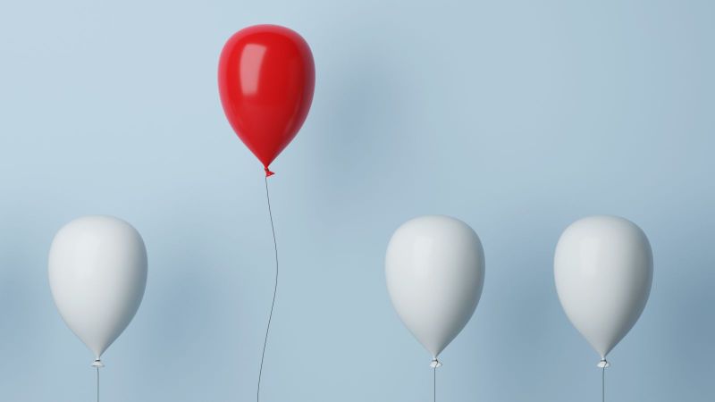 Ein roter Luftballon hebt sich von mehreren weißen Luftballons ab.